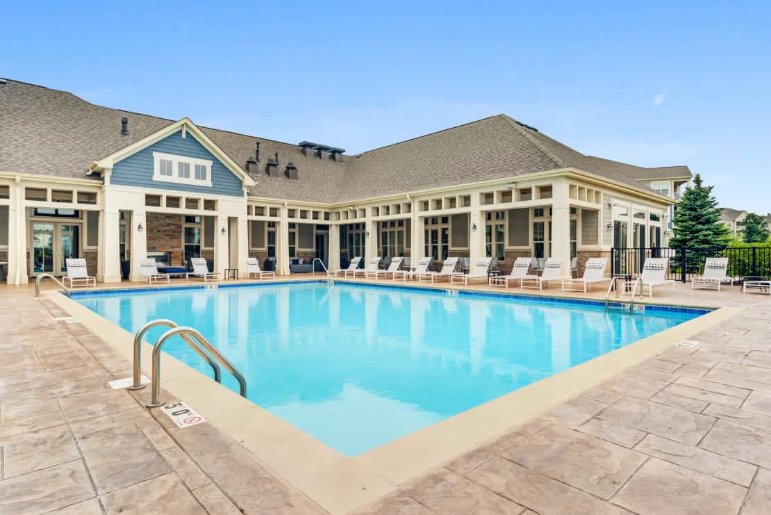 Resort-style swimming pool with clear blue water, concrete deck, white lounge chairs, and a large cream and blue clubhouse with multiple windows and covered porches.