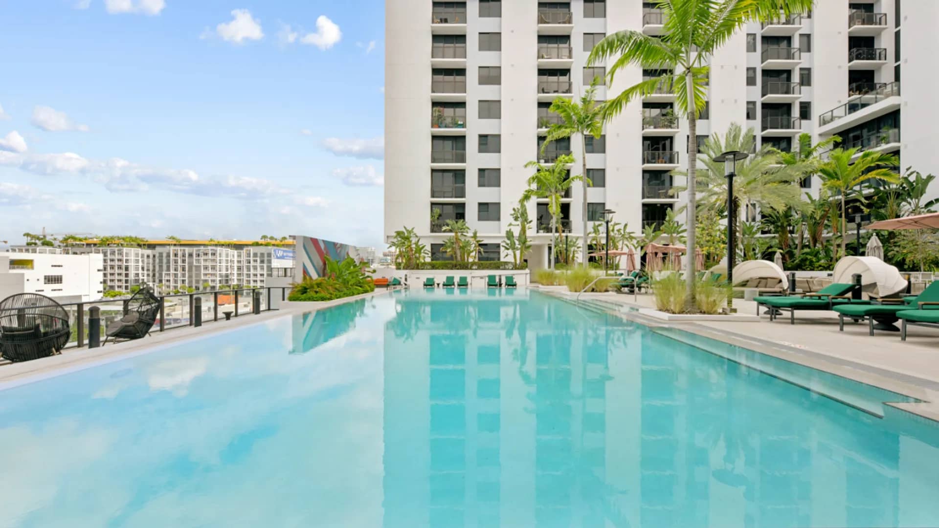 Rooftop swimming pool with turquoise water, lounge chairs, umbrellas, tropical plants, and modern residential tower in background.