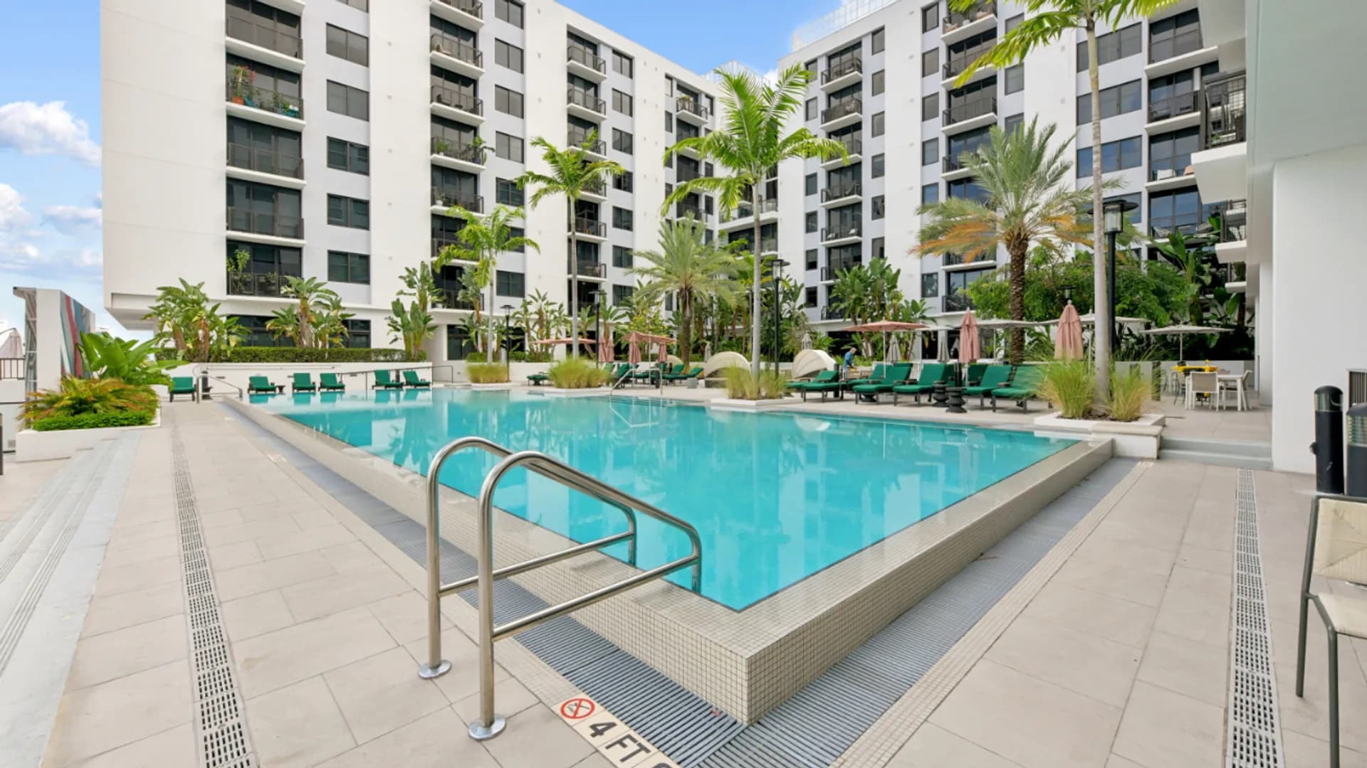 Resort-style swimming pool with metal handrails, surrounded by palm trees, lounge chairs, and white multi-story apartment buildings under clear sky.