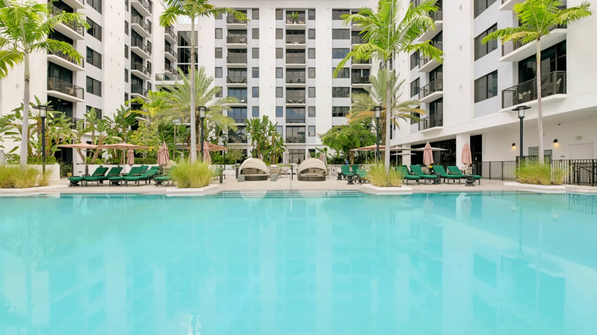 Resort pool with turquoise water, green lounge chairs, palm trees, and white multi-story residential buildings surrounding a courtyard.