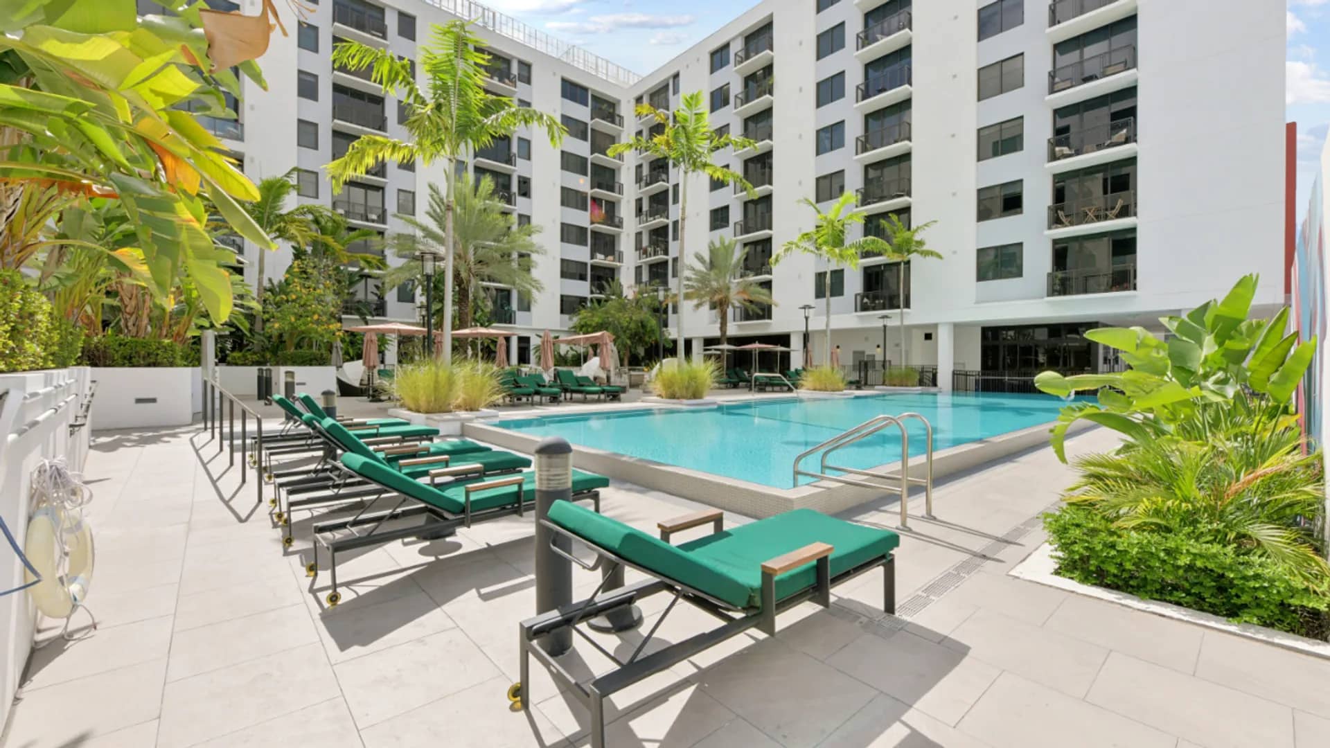 Resort pool with turquoise water, green lounge chairs, palm trees, and white apartment buildings in background.