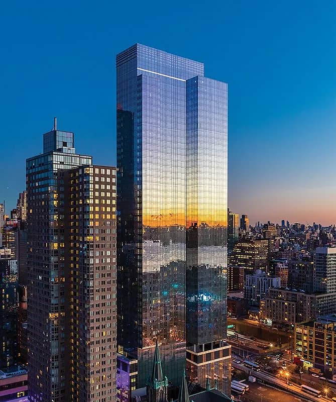 Modern glass high-rise tower with reflective facade showing sunset and Manhattan skyline, surrounded by neighboring buildings at dusk.