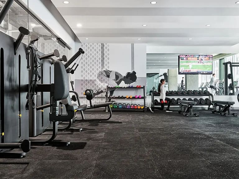 Modern gym with cardio equipment on left, dumbbell rack in center, and resident exercising on equipment, with recessed ceiling lights and dark flooring.