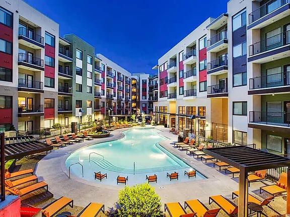 Illuminated resort pool at dusk surrounded by multi-story apartment buildings with red, green, and blue facades, lounge chairs, and landscaping.
