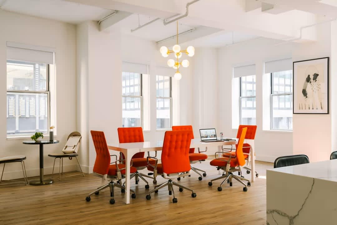 Open office space with white round table, red office chairs, hardwood floors, multiple windows with city views, and brass pendant light fixture.