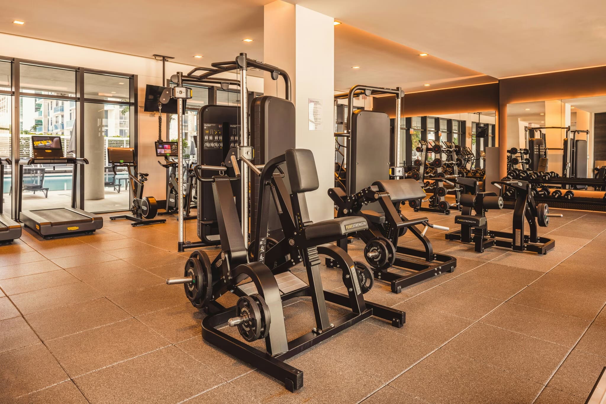 Modern gym with stationary bikes, weight machines, and strength equipment on cork flooring, large windows with city views.