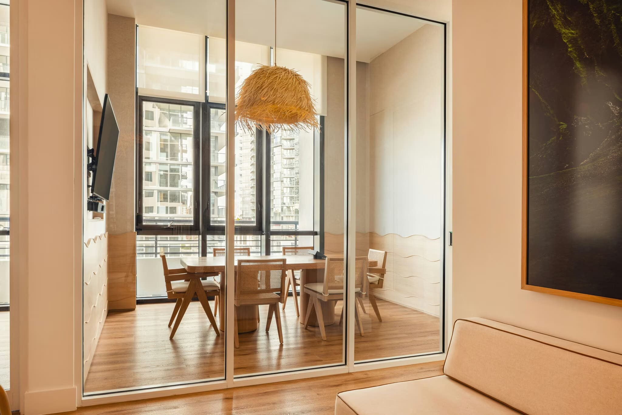 Dining room with wooden table, rattan pendant light, glass sliding doors, and high-rise city views through floor-to-ceiling windows.