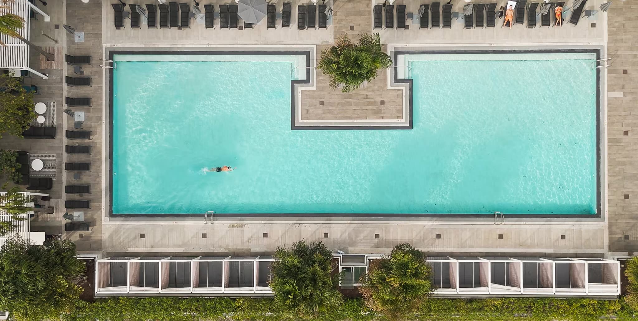 Overhead view of a large turquoise swimming pool with sun loungers, surrounded by brick buildings and landscaping.
