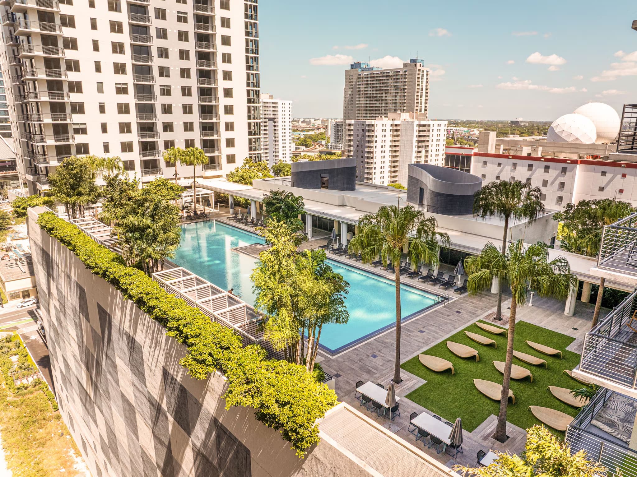 Elevated view of rectangular swimming pool surrounded by palm trees, lounge chairs, and manicured lawn with high-rise residential buildings in background.