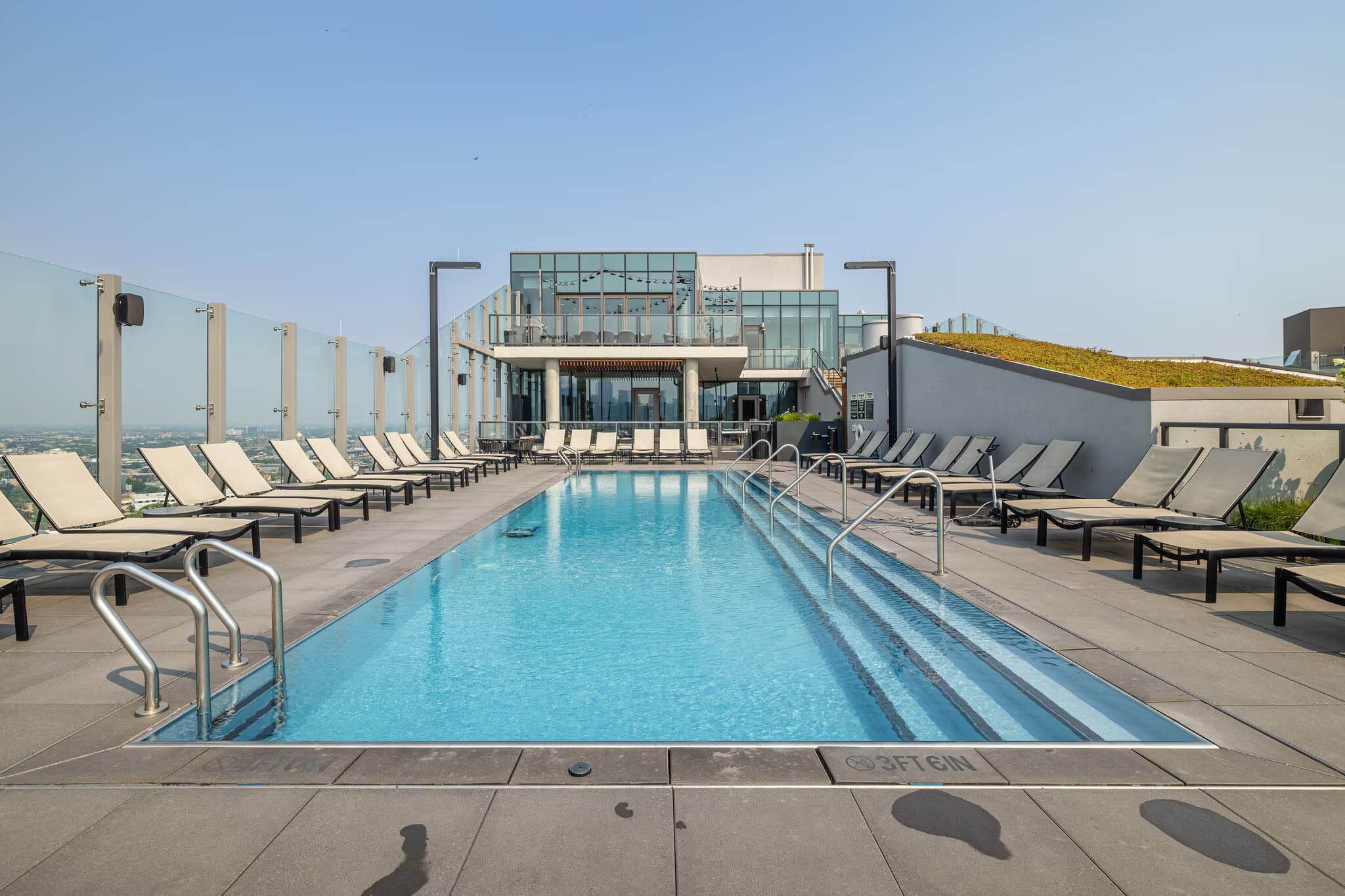 Rooftop swimming pool with lounge chairs, metal railings, and modern building with glass windows overlooking cityscape.