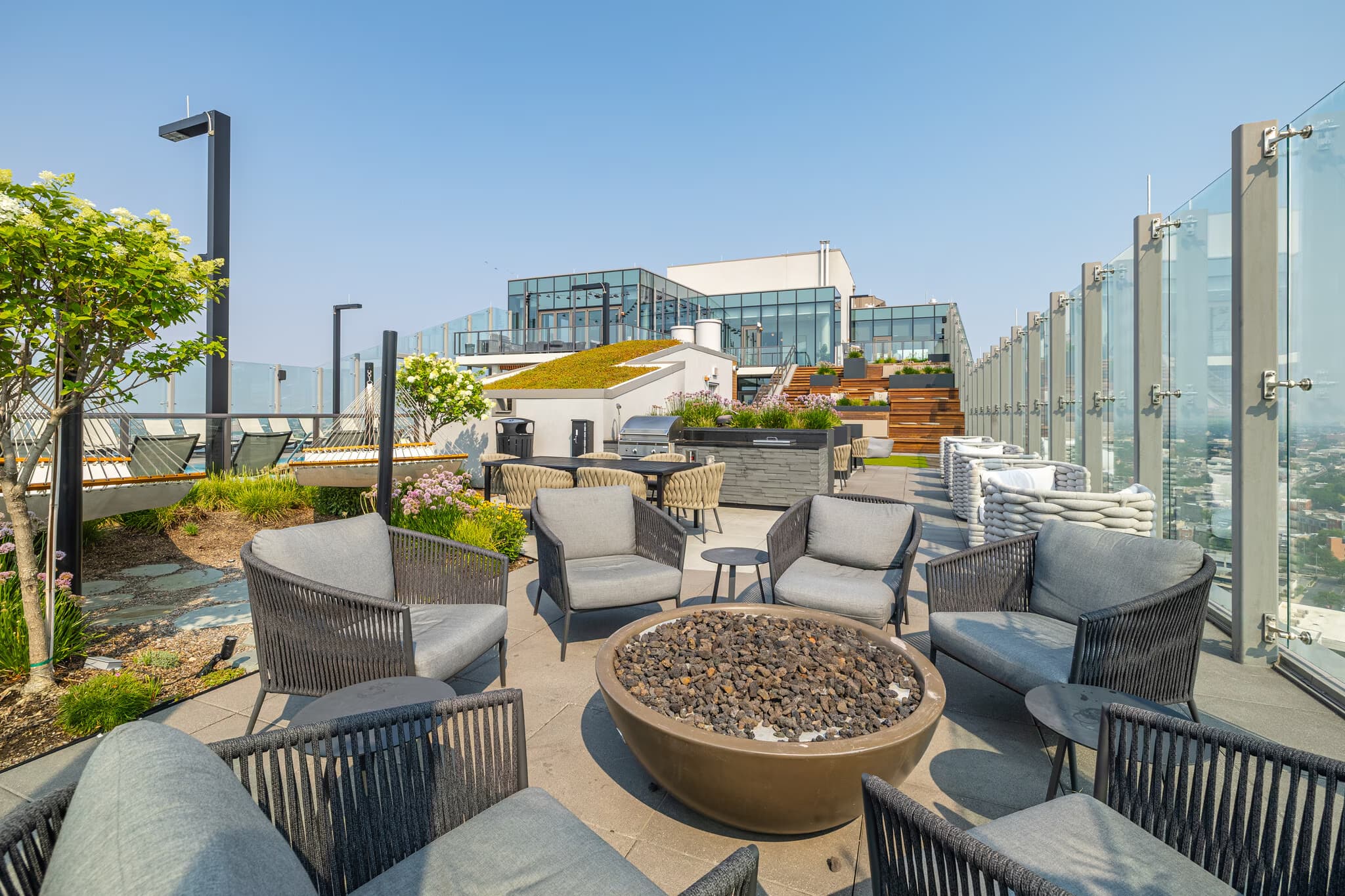 Rooftop terrace with woven lounge chairs arranged around a circular fire pit, green plantings, and glass railings overlooking the city.