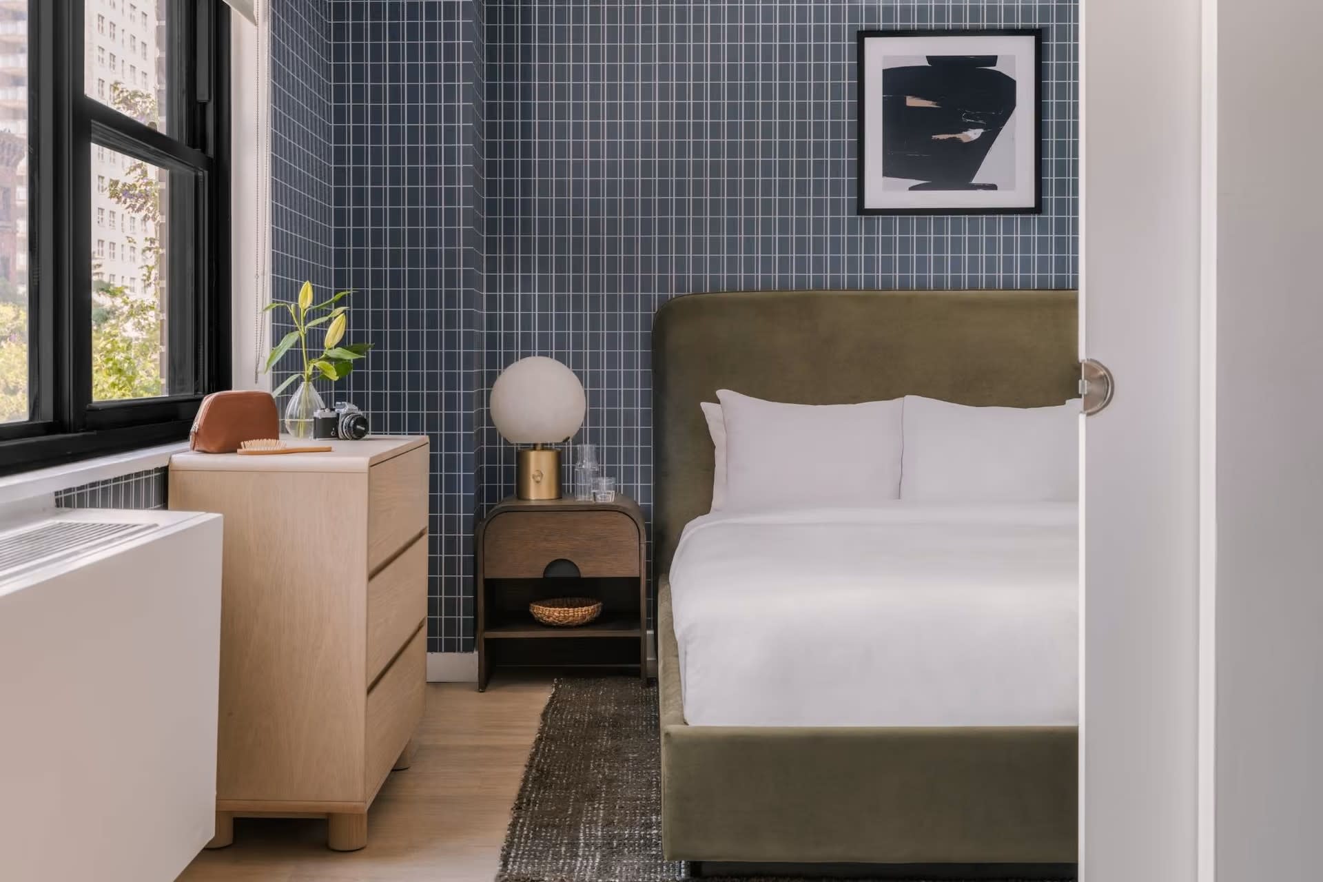 Bedroom with olive-green upholstered bed, white bedding, blue grid accent wall, wooden nightstand, and large window with city views.