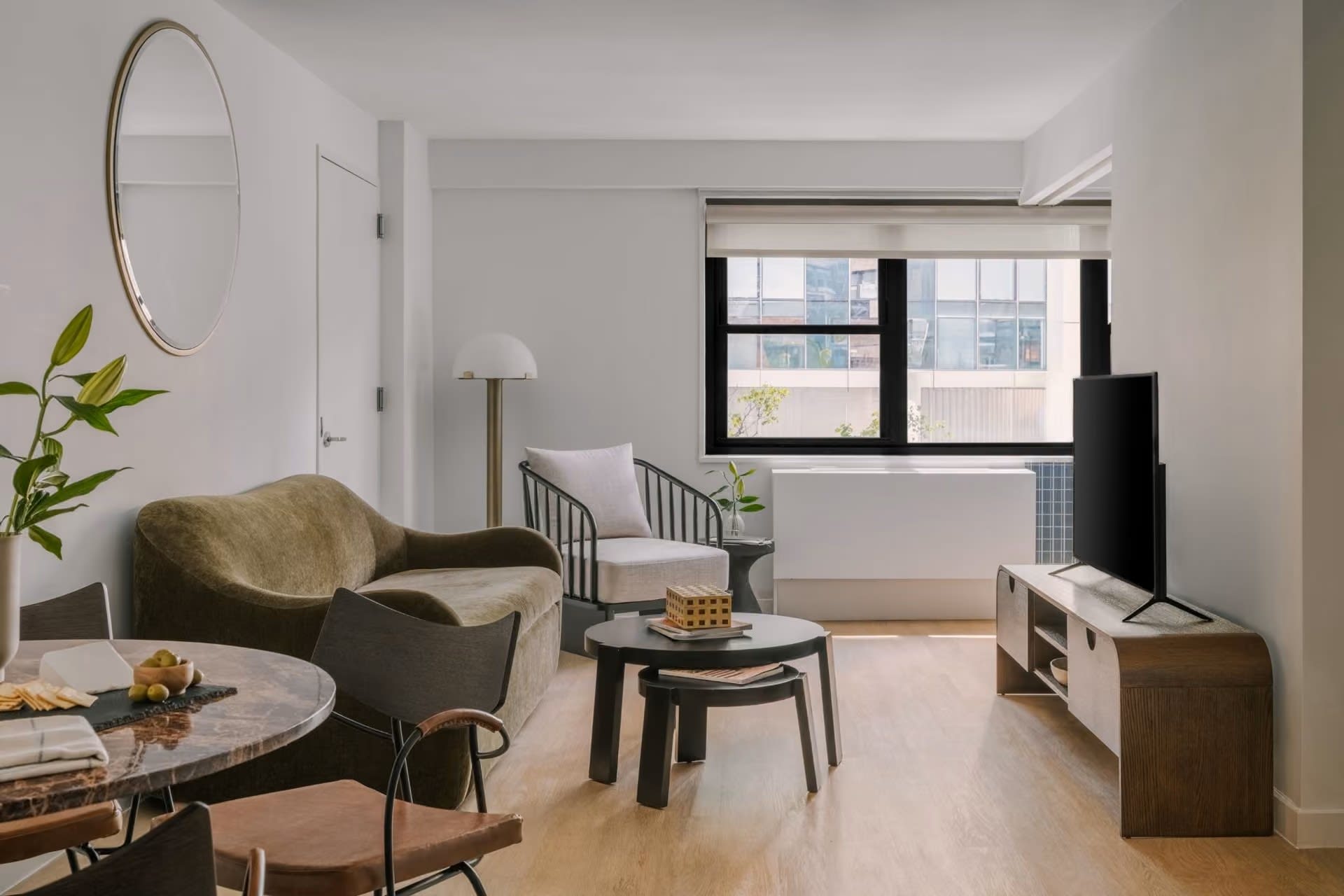 Modern living room with olive-green armchair, black coffee table, wooden TV stand, and floor-to-ceiling windows with city views.