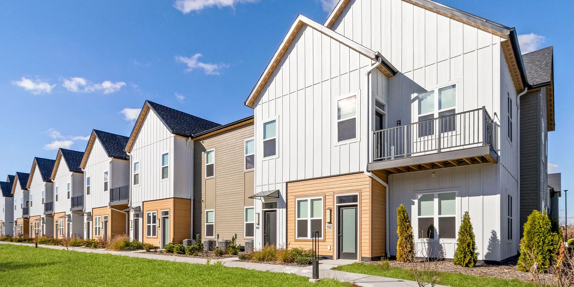 Row of contemporary townhomes with white vertical siding, wood accents, second-floor balconies, and manicured landscaping.
