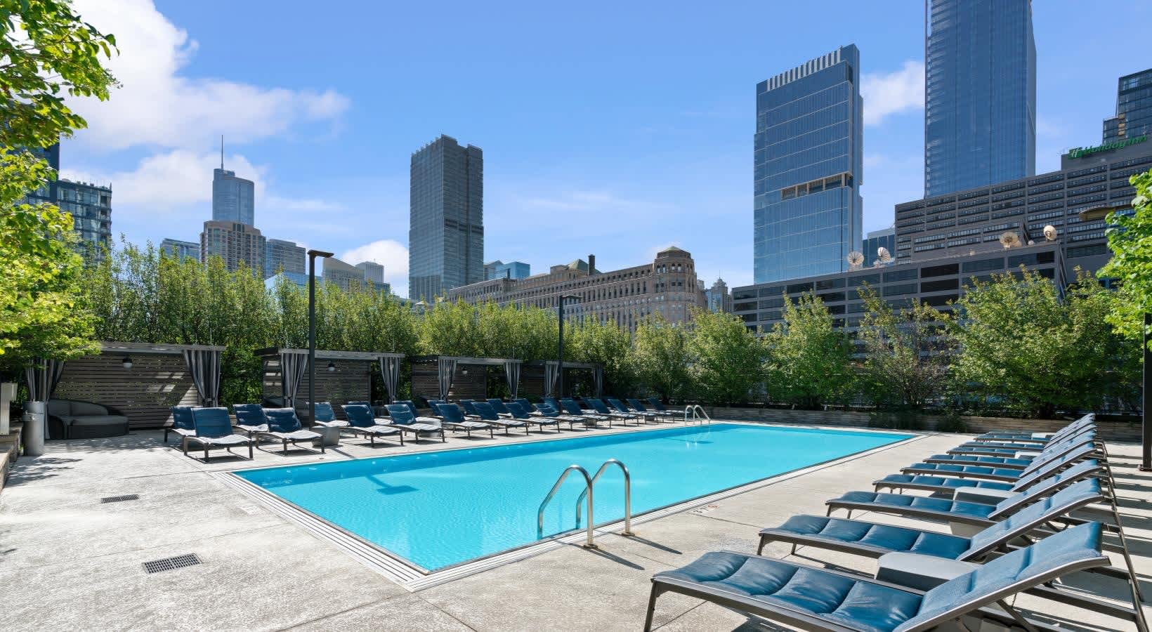 Rooftop swimming pool with blue lounge chairs, metal railings, and Chicago skyline with tall buildings in background.