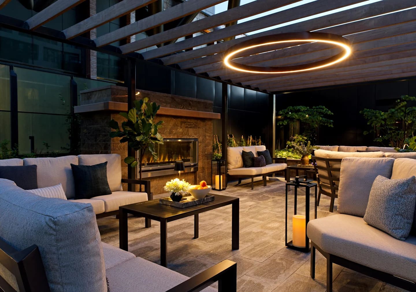 Rooftop lounge with cream upholstered seating, black tables, stone fireplace, and circular gold ceiling light fixture at night.