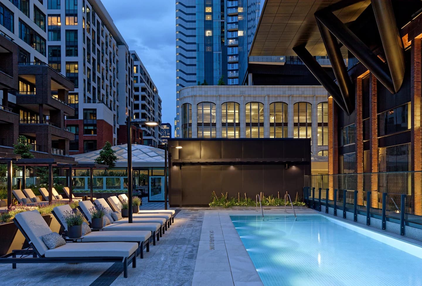 Outdoor swimming pool at dusk with lounge chairs, surrounded by contemporary apartment buildings and a black accent wall.