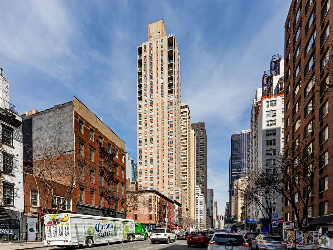 Street view of tall residential building with tan brick facade among surrounding Manhattan buildings and parked cars.
