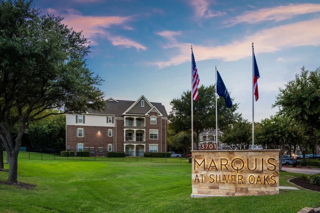 Brick colonial apartment building with white trim, green lawn, mature oak trees, and Marquis at Silver Oaks monument sign with three flags.