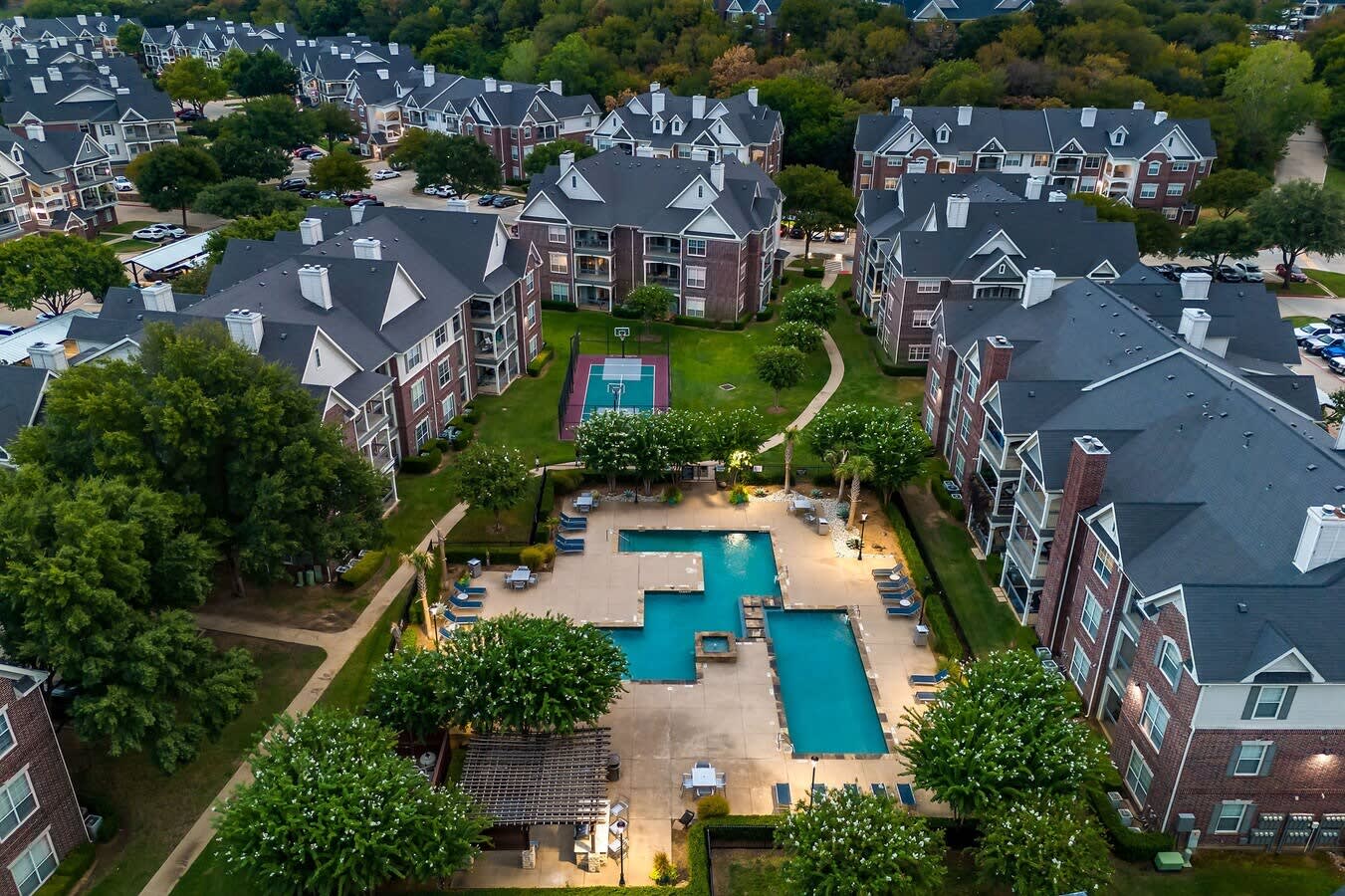 Aerial view of apartment complex with turquoise swimming pools, basketball court, and brick buildings with dark roofs surrounded by mature trees.
