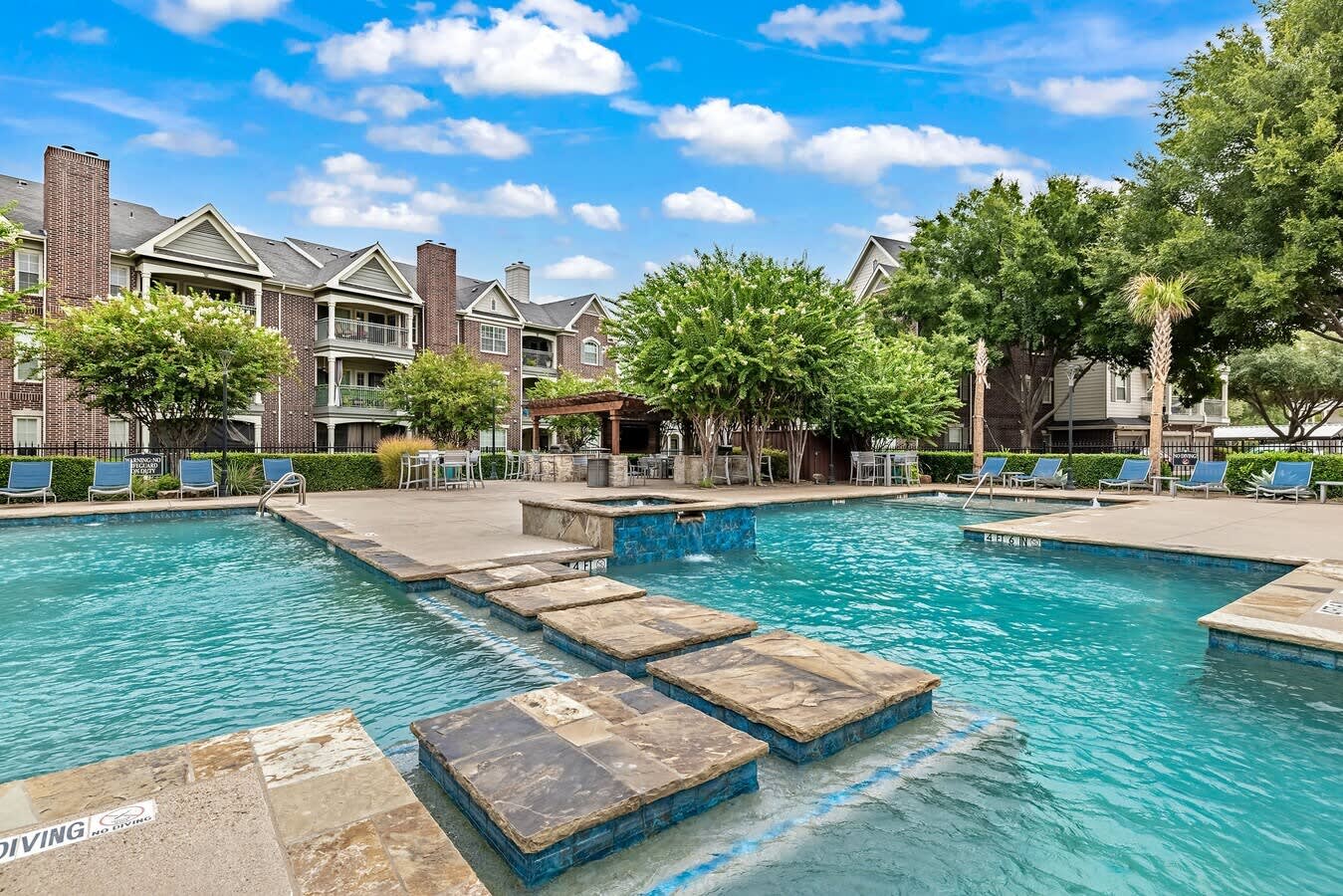 Resort-style swimming pool with turquoise water, stepping stones, lounge chairs, and multi-story brick apartment buildings with mature trees in background.