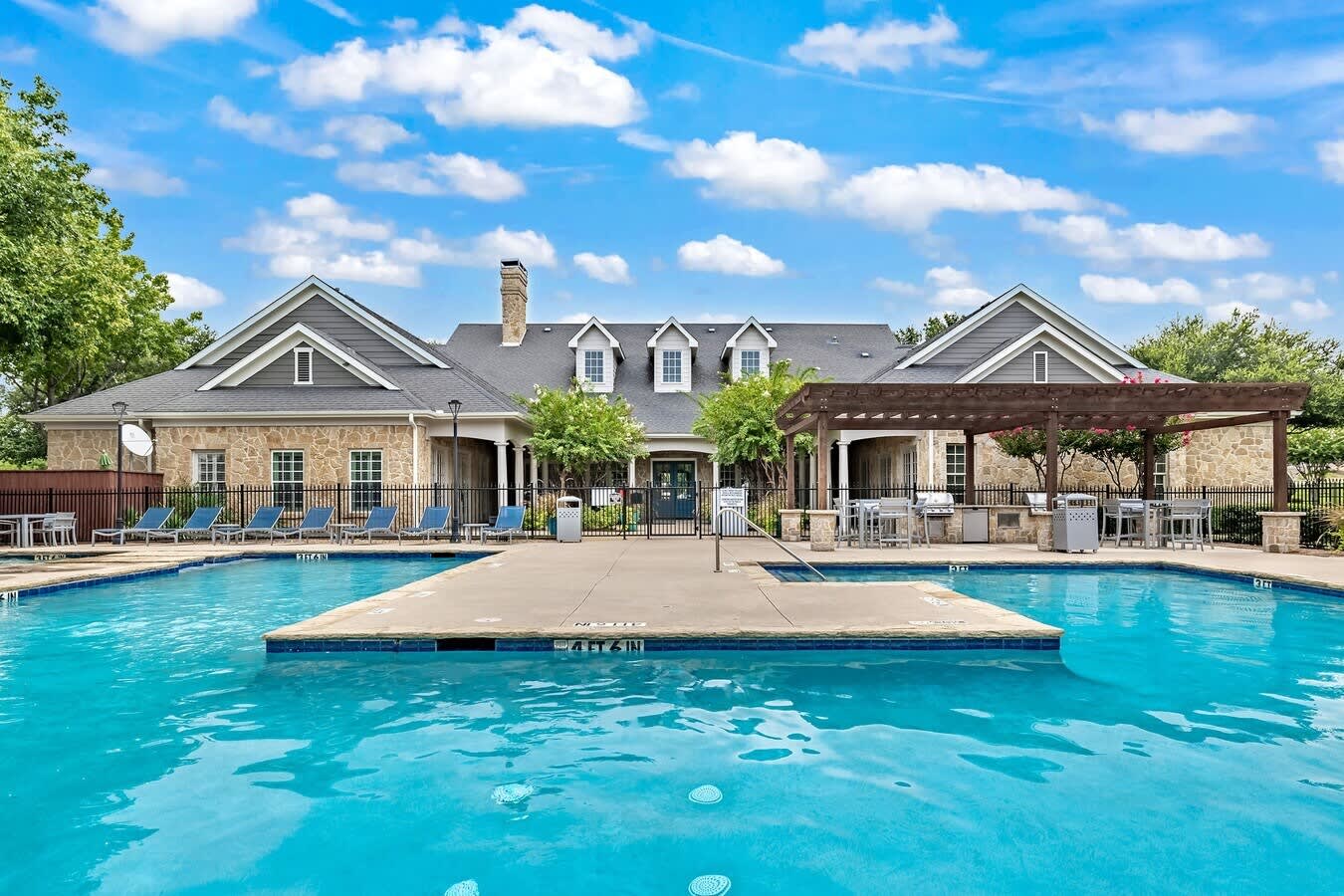 Two bright blue pools with diving board facing a stone and siding clubhouse with white columns, pergola, and lounge chairs under clear sky.