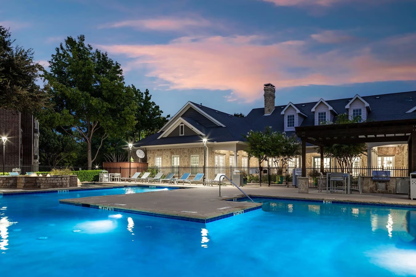 Lit swimming pools with lounge chairs, stone clubhouse with pergola, and residential buildings at twilight.