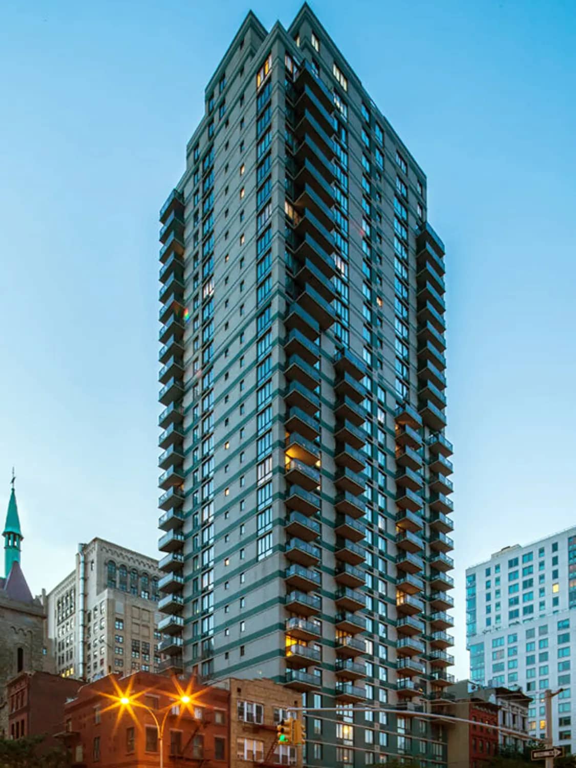 Tall residential tower with teal-gray facade, stacked balconies, and lit windows at dusk surrounded by historic buildings.