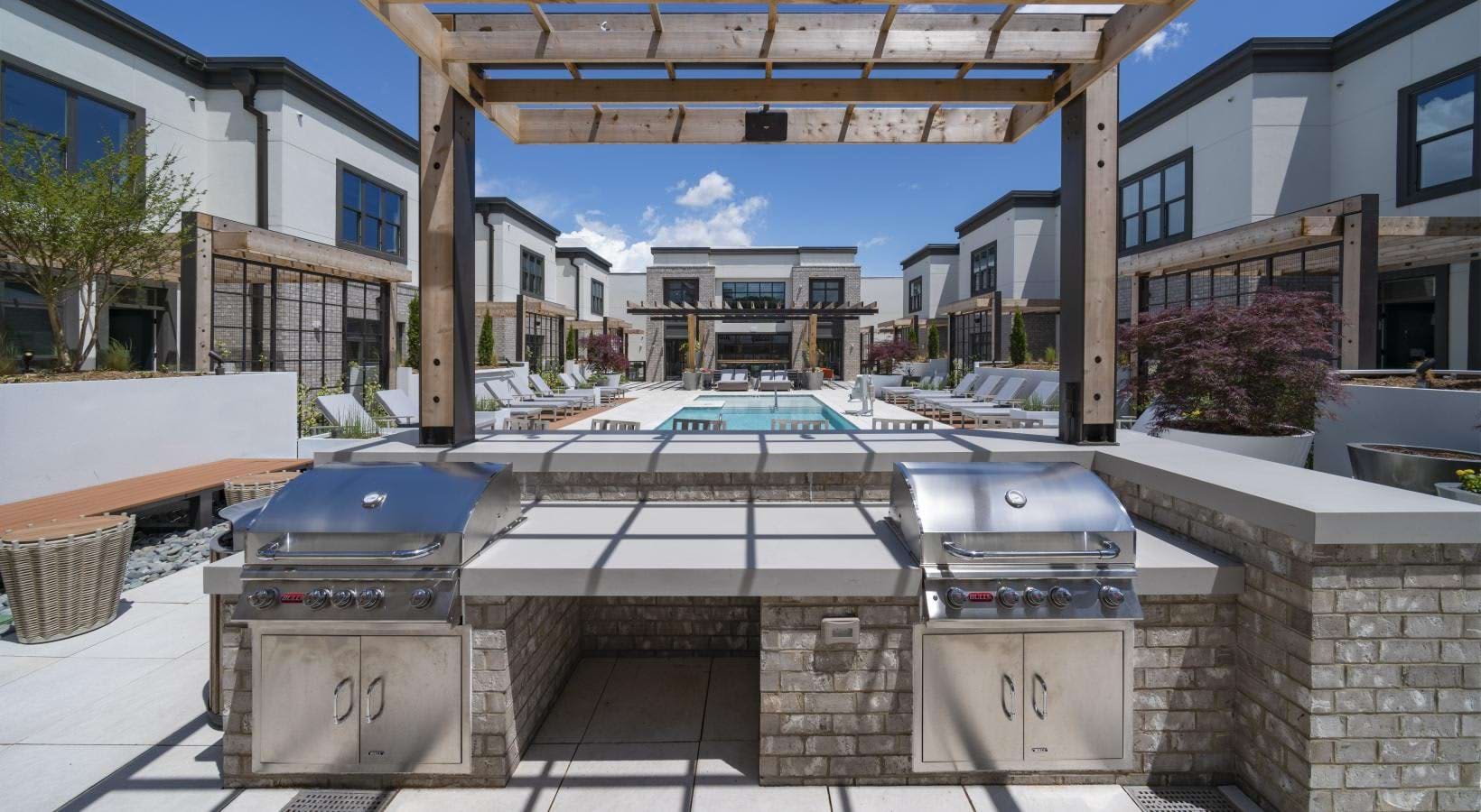 Outdoor courtyard with two stainless steel grills, stone counters, pool, lounge seating, and white modern townhomes under pergola.