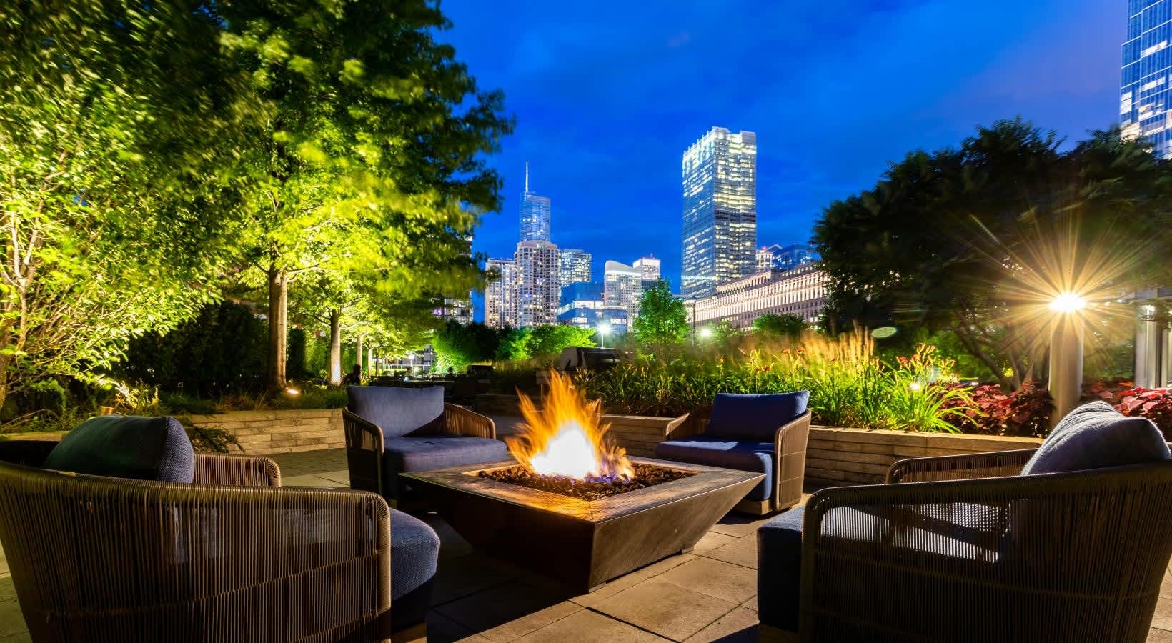 Rooftop terrace at dusk with lit fire pit, woven lounge chairs, planted containers, and Chicago skyline backdrop.