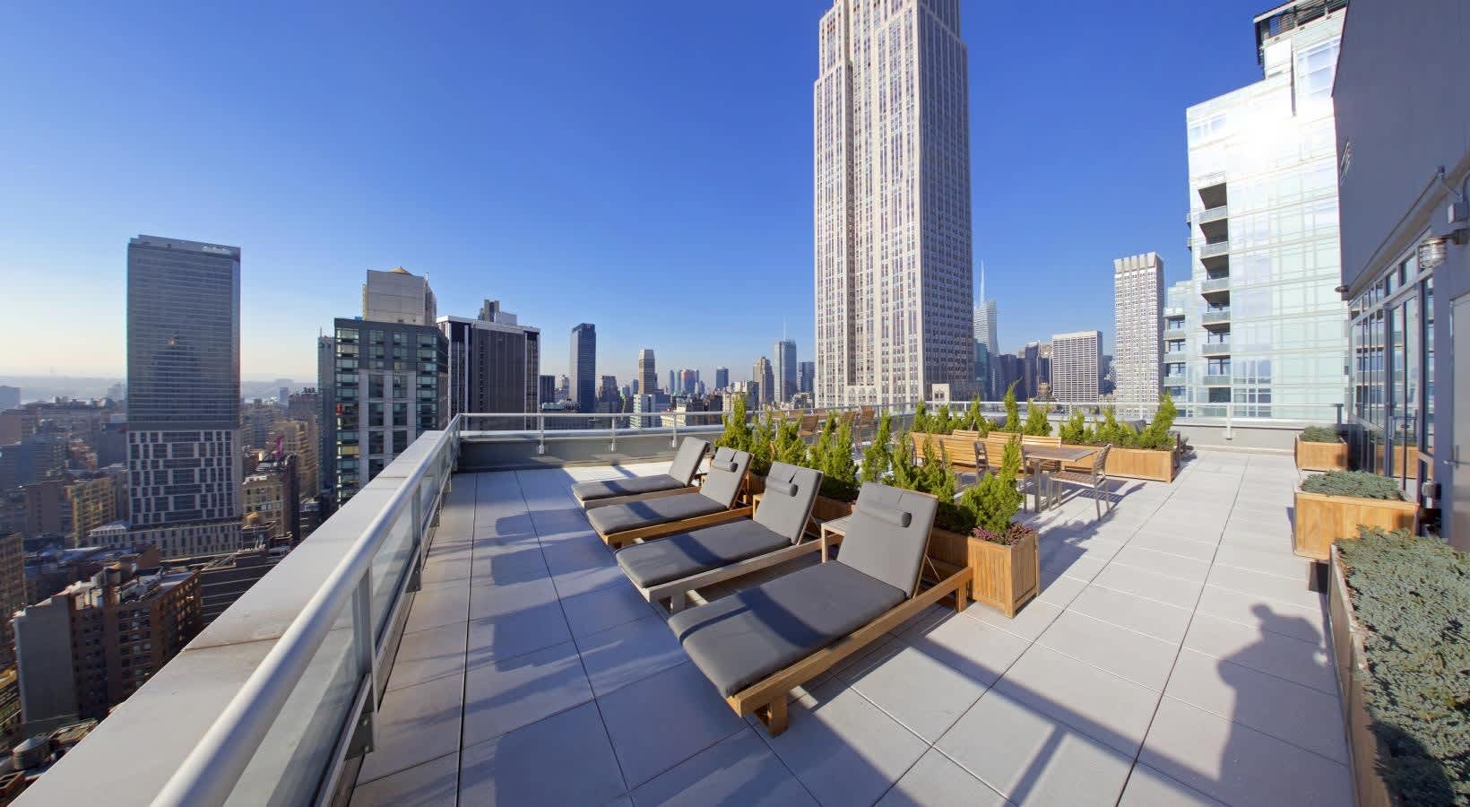 Rooftop terrace with wooden lounge chairs, potted plants, and Manhattan skyline views under clear blue sky.
