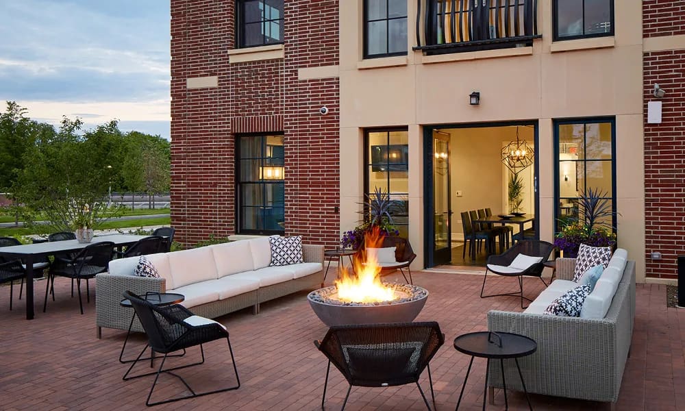 Outdoor courtyard with lit fire pit, white sectional sofas, black chairs, and brick building facade at dusk.