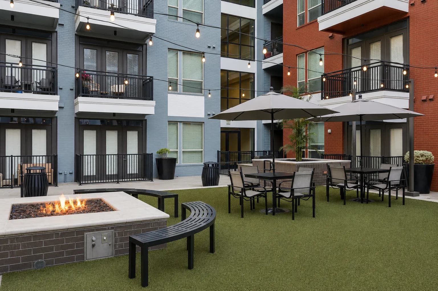 Outdoor courtyard with fire pit, dining tables under umbrellas, and artificial turf surrounded by multi-story residential buildings with balconies.