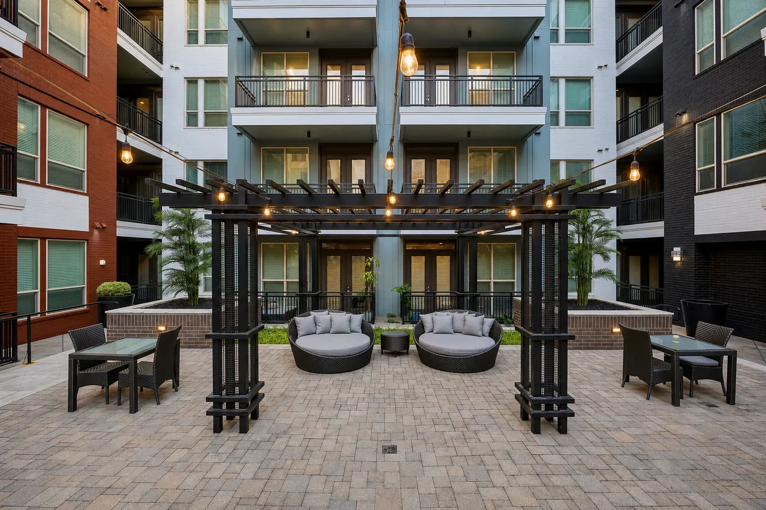 Courtyard with two round gray lounge chairs under black pergola with hanging lights, surrounded by multi-story apartment buildings with balconies.