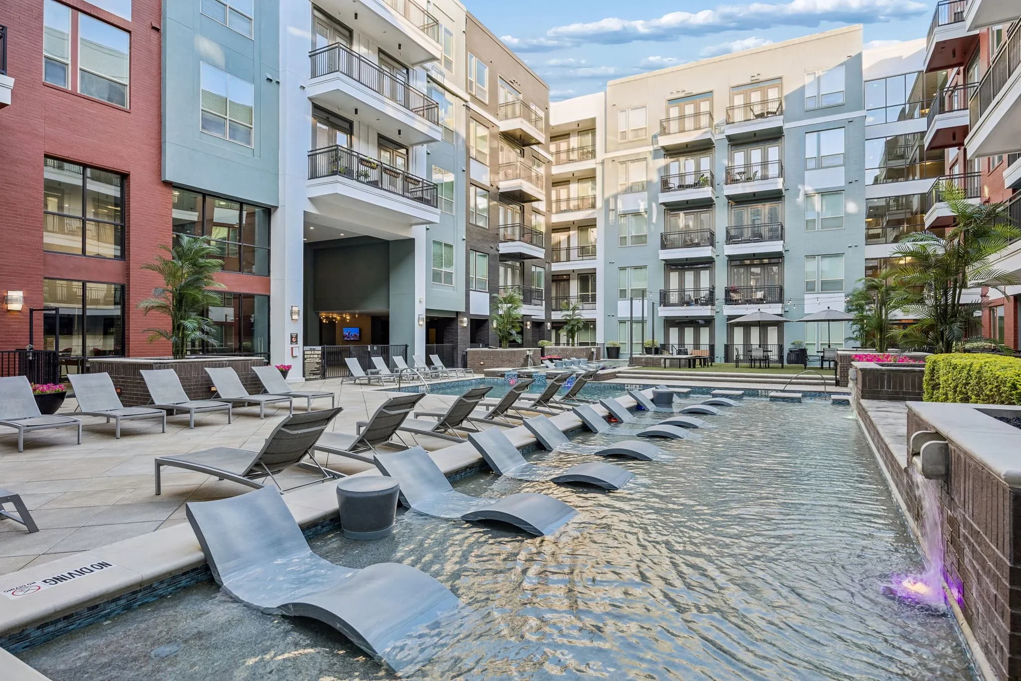 Resort pool with lounge chairs in shallow water, surrounded by red, blue, and tan apartment buildings in a courtyard setting.