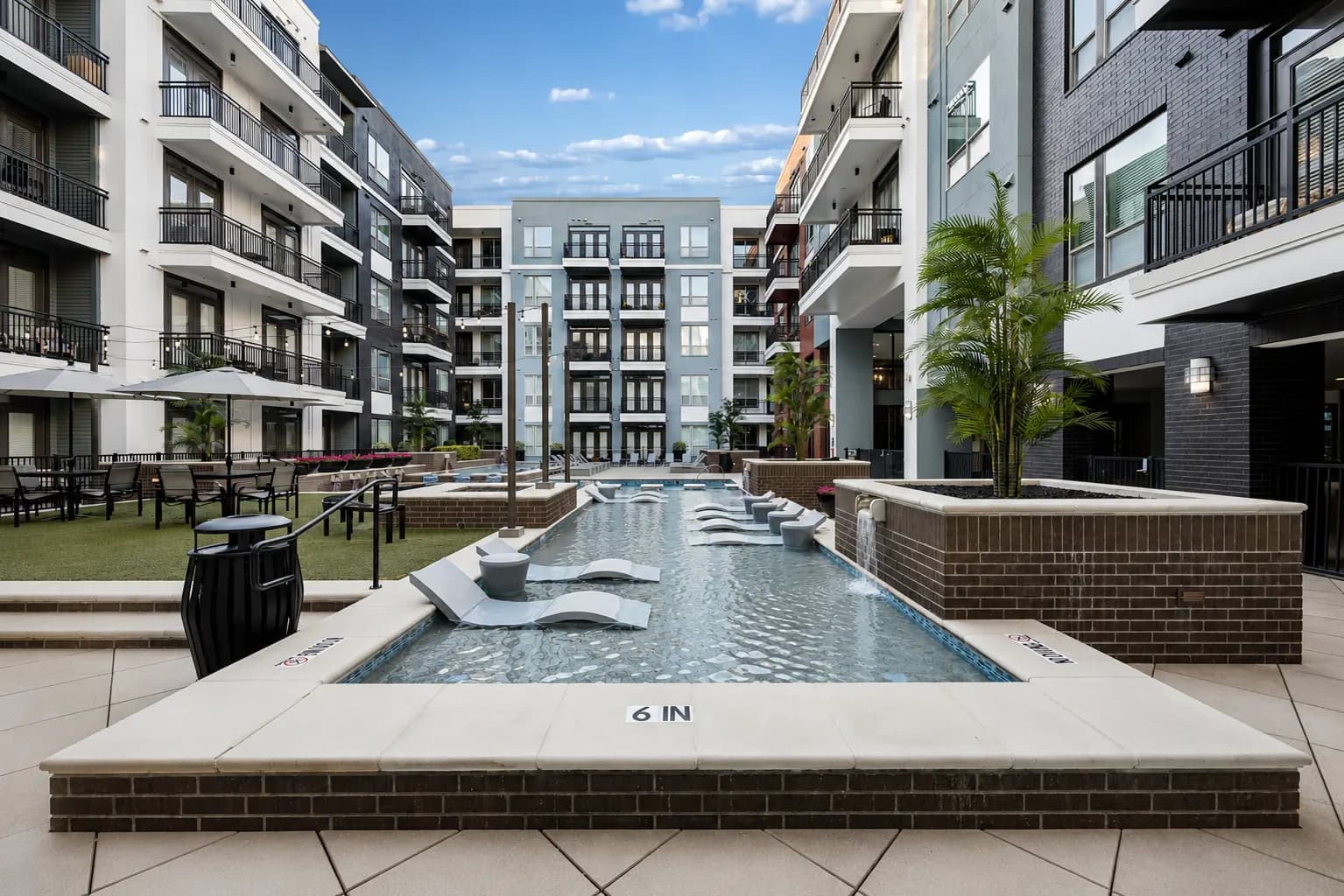 Resort-style pool with white lounge chairs in a courtyard surrounded by multi-story apartment buildings with balconies and palm trees.