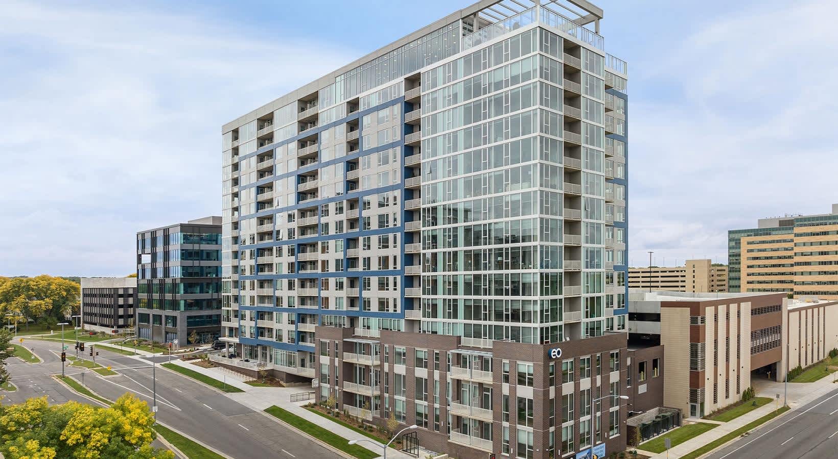 Contemporary mid-rise residential building with glass and brick facade, ground-floor retail, tree-lined streets, and adjacent commercial buildings.