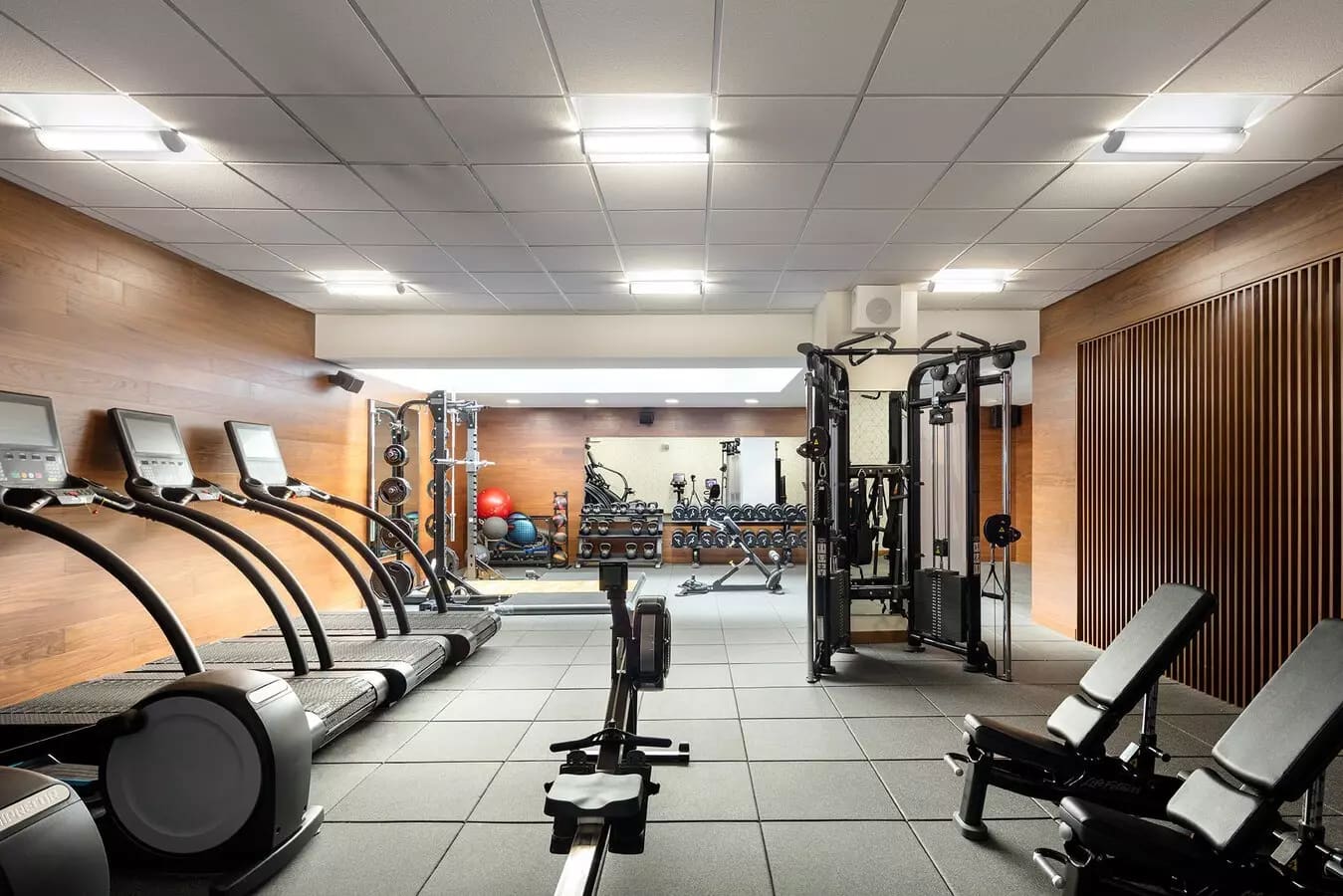 Modern gym with treadmills on left, stationary bikes in center, weight machines on right, and mirrored walls under bright ceiling lights.
