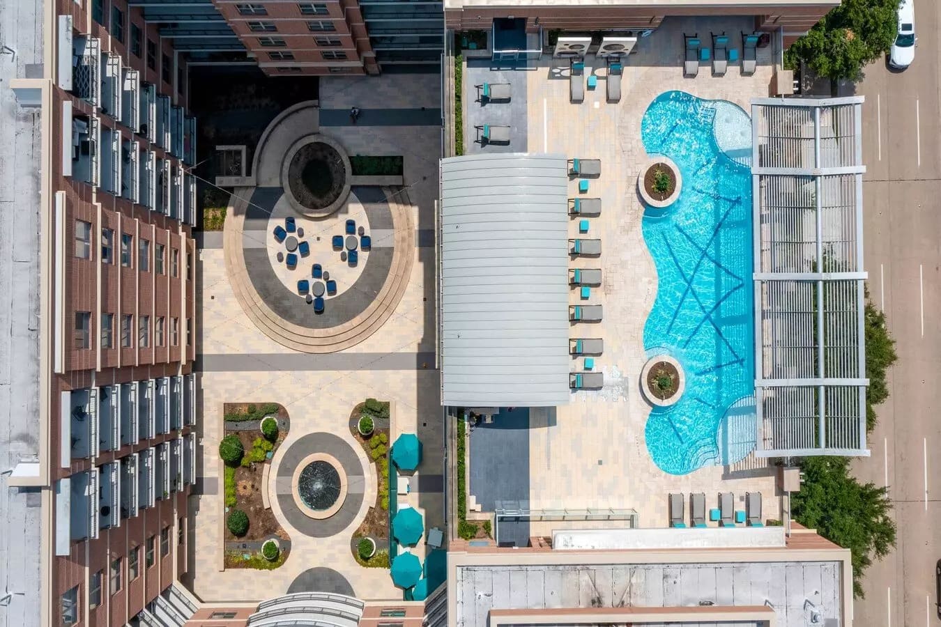 Overhead view of turquoise pool with white shade structures, lounge chairs, and circular courtyard with seating areas surrounded by brick residential buildings.