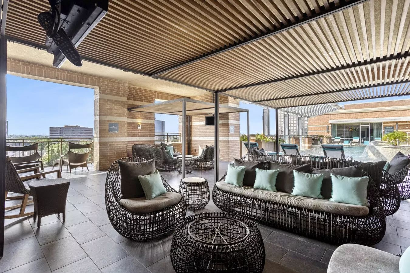 Rooftop terrace with black woven lounge chairs, sage green pillows, slatted pergola overhead, and Houston skyline views.