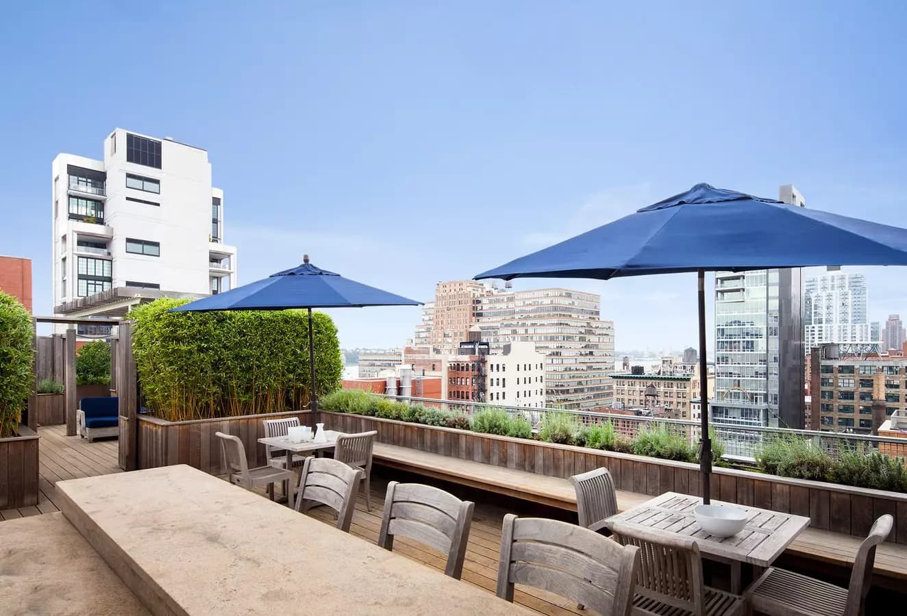 Rooftop terrace with wooden dining tables, gray chairs, blue umbrellas, planters, and panoramic Manhattan skyline views.