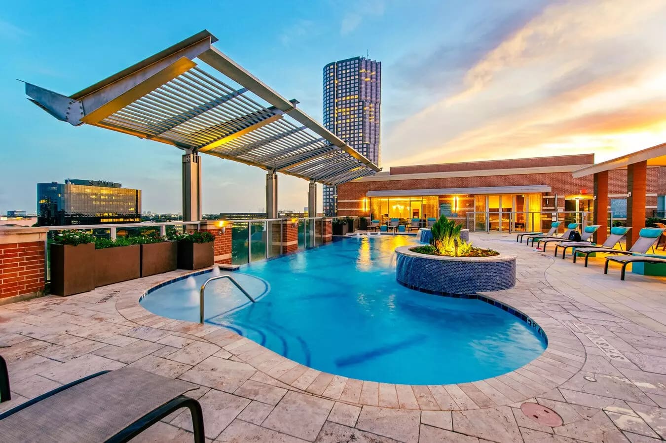 Rooftop pool area at dusk with blue water, lounge chairs, pergola, fire pit, and Houston skyline in background.