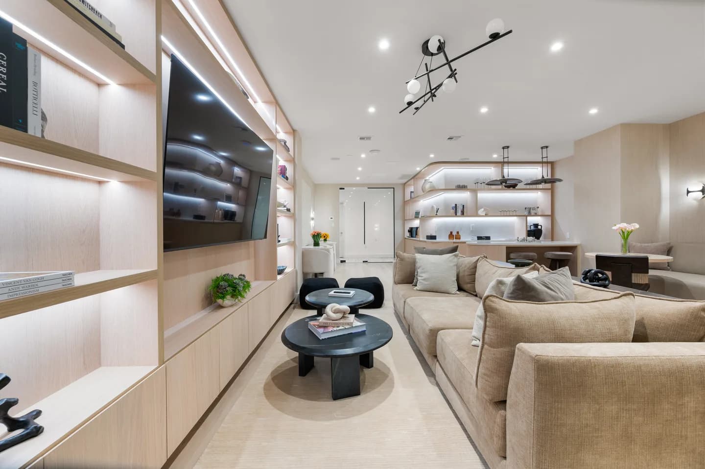 Contemporary living room with wall-mounted TV, beige sectional sofa, black round coffee table, and floor-to-ceiling shelving with recessed lighting.