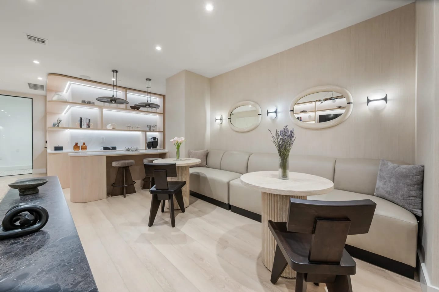 Open-concept lounge with cream sectional sofa, round white tables, wood bar with stools, and illuminated shelving.