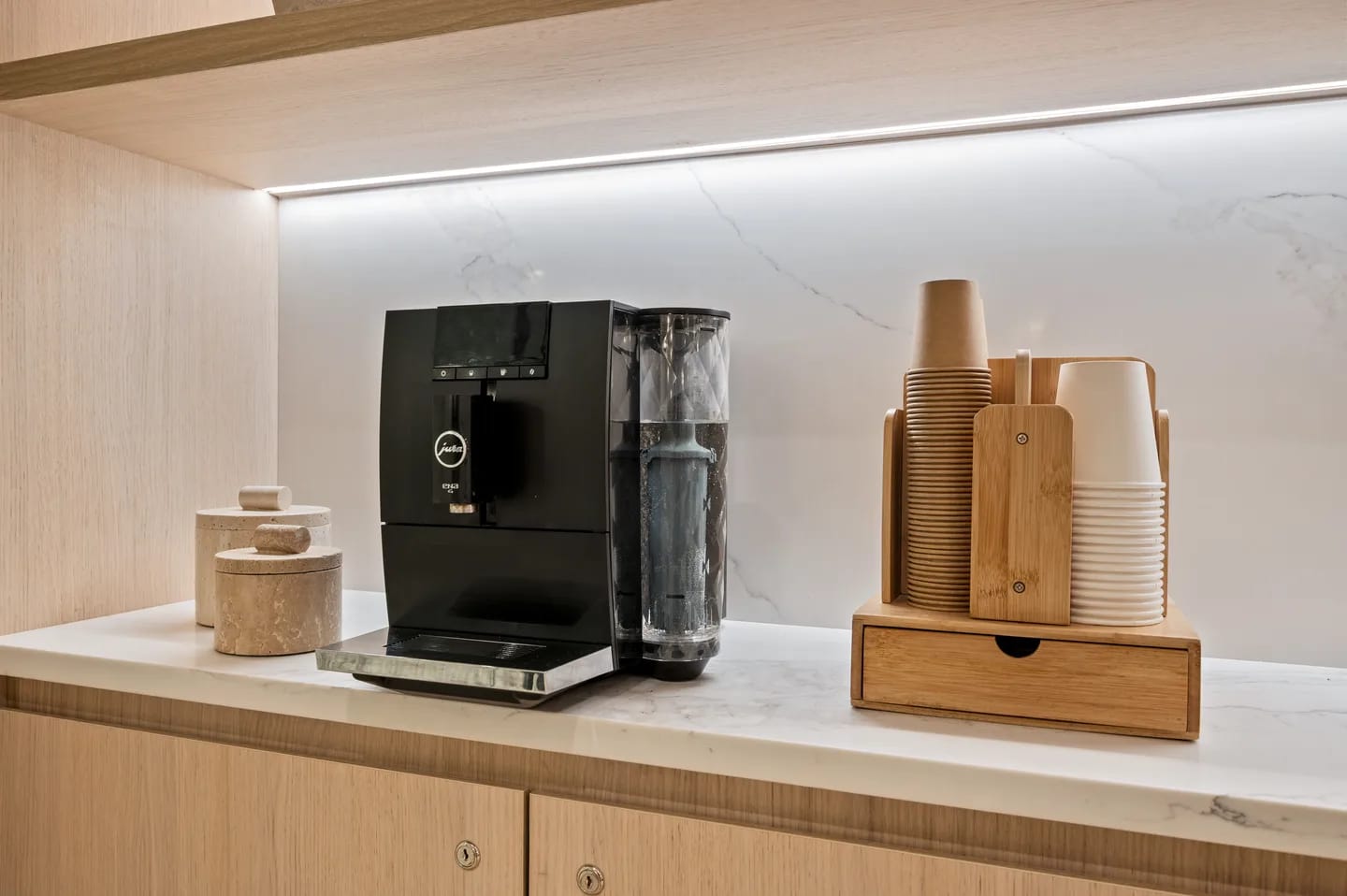 Kitchenette with black automatic coffee machine, water dispenser, wooden cup holder with stacked cups, and light wood cabinetry.