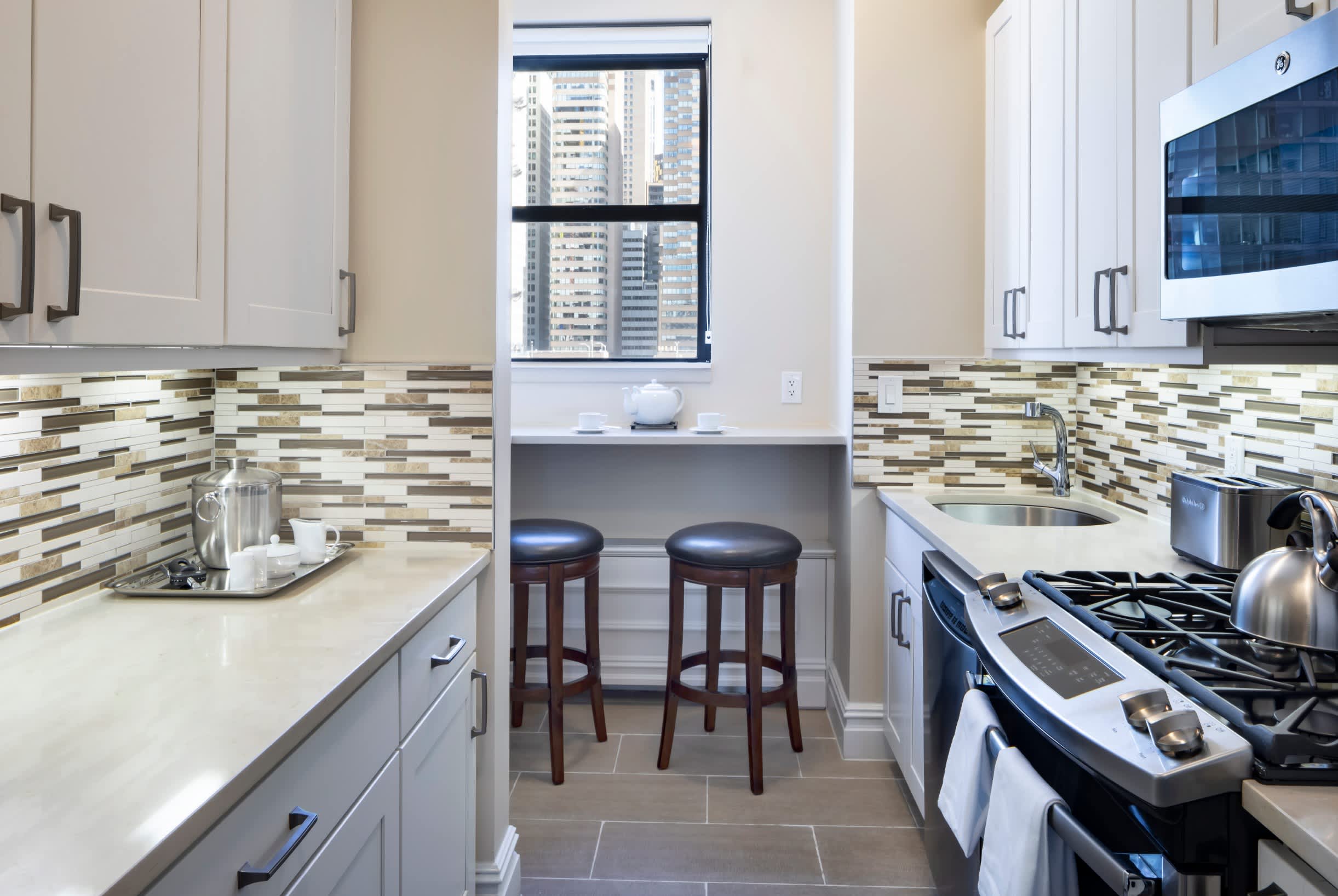 Modern kitchen with white cabinets, multicolor tile backsplash, gas range, and two bar stools at window counter overlooking city buildings.