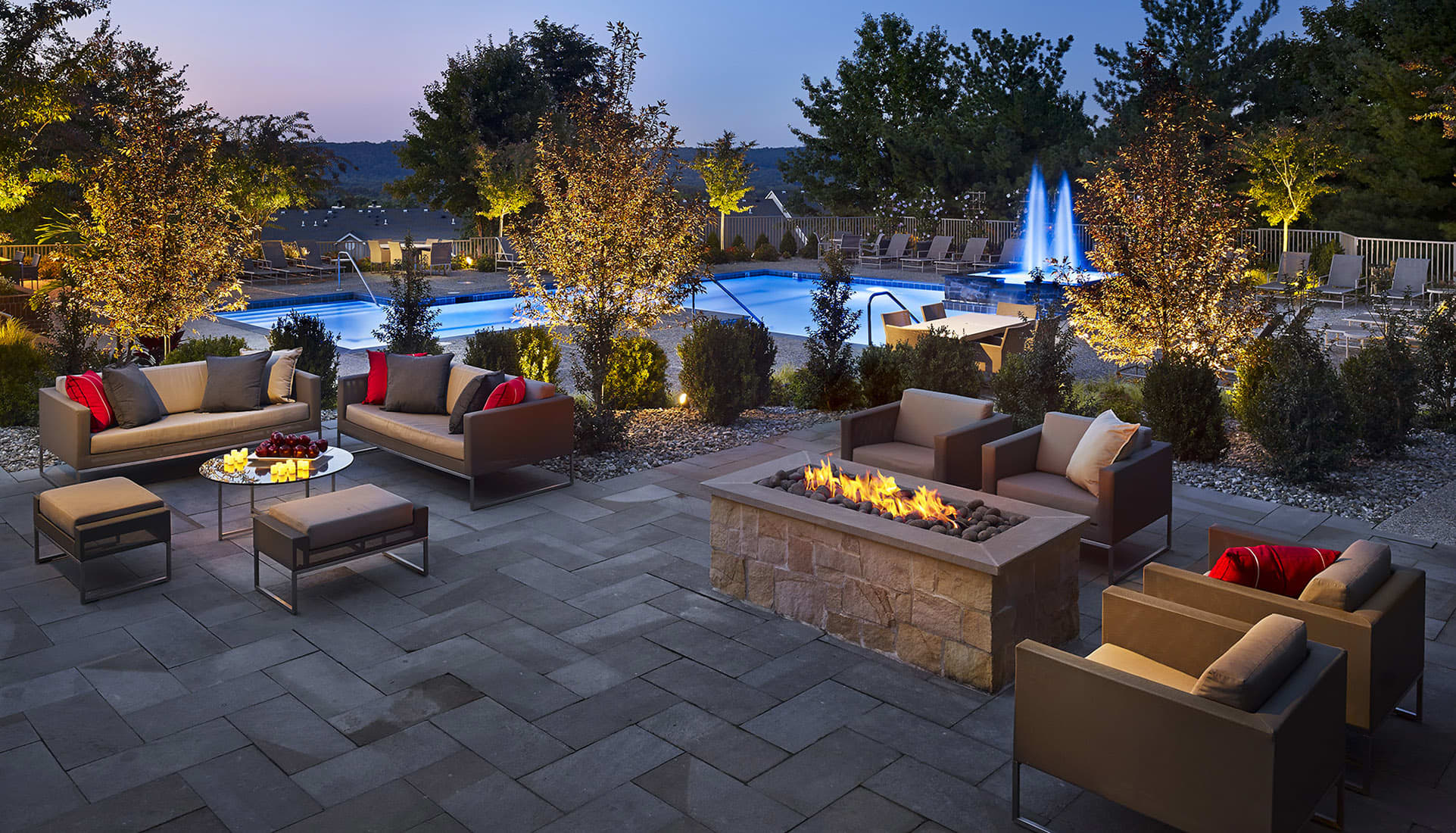 Outdoor patio at dusk with modern seating, linear fireplace, lit trees, and illuminated pool with fountain in background.