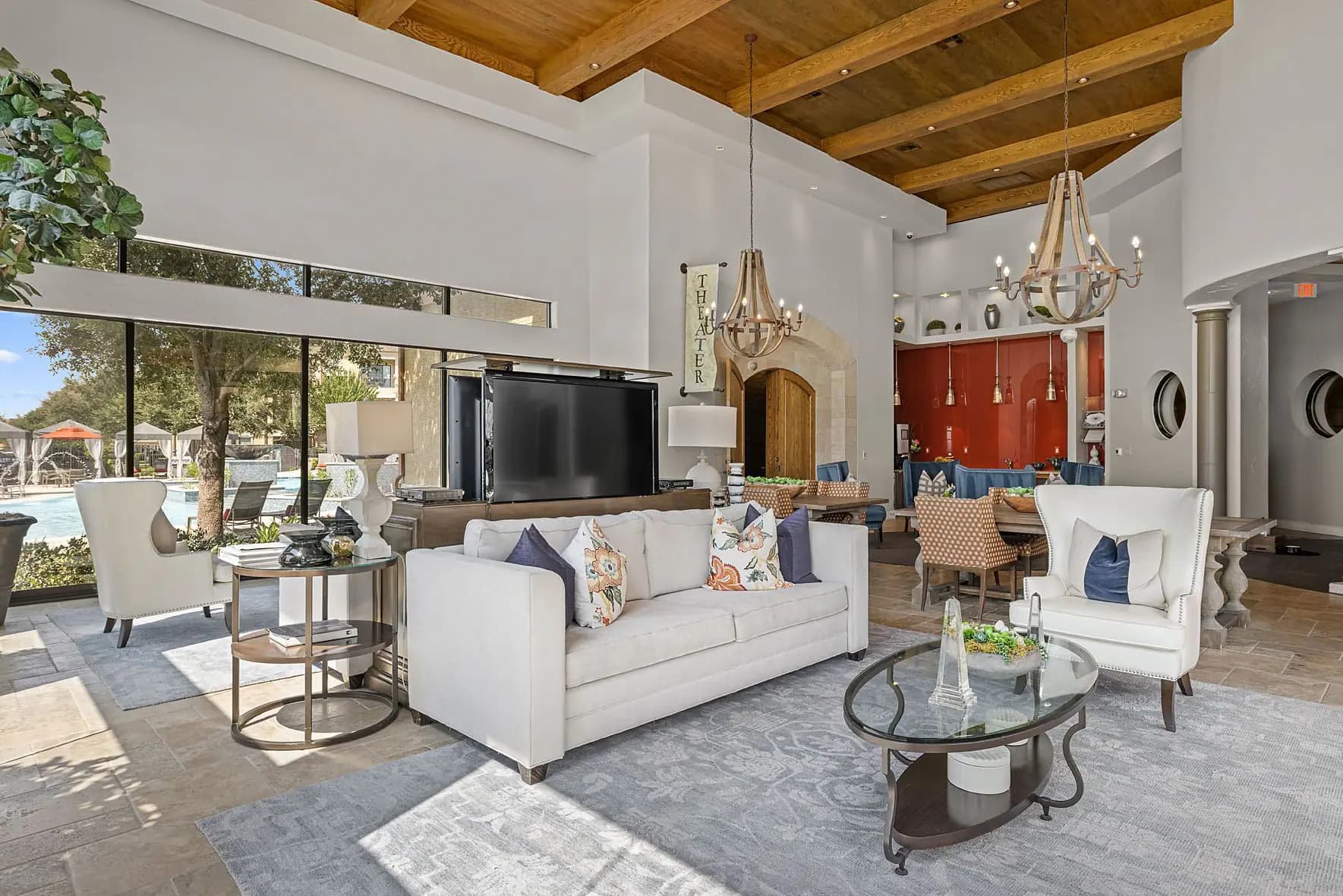 Living room with cream sofa, exposed wooden ceiling beams, pendant chandeliers, and floor-to-ceiling windows with pool view.
