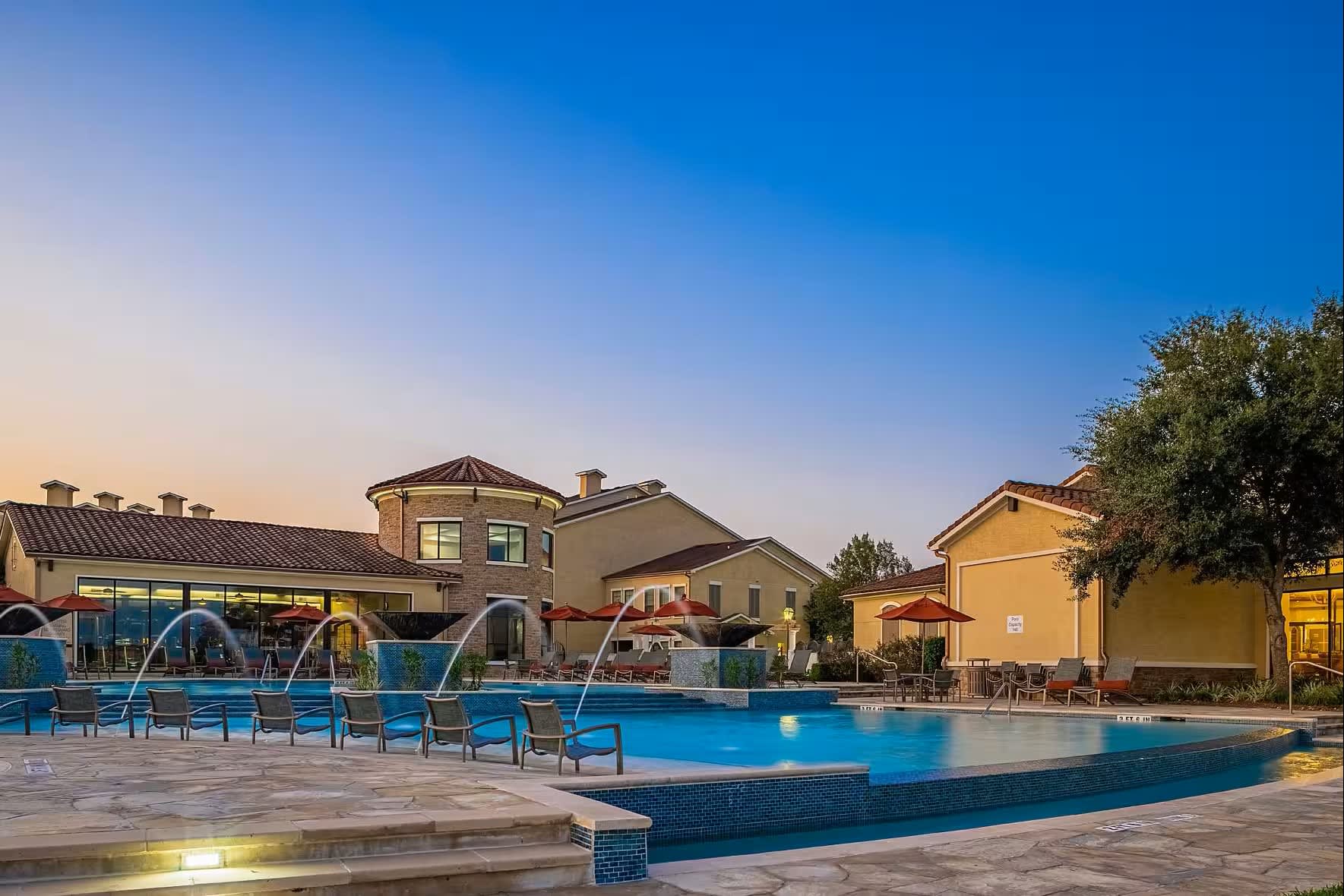 Resort-style swimming pool with blue lounge chairs, fountains, and surrounding residential buildings at twilight.