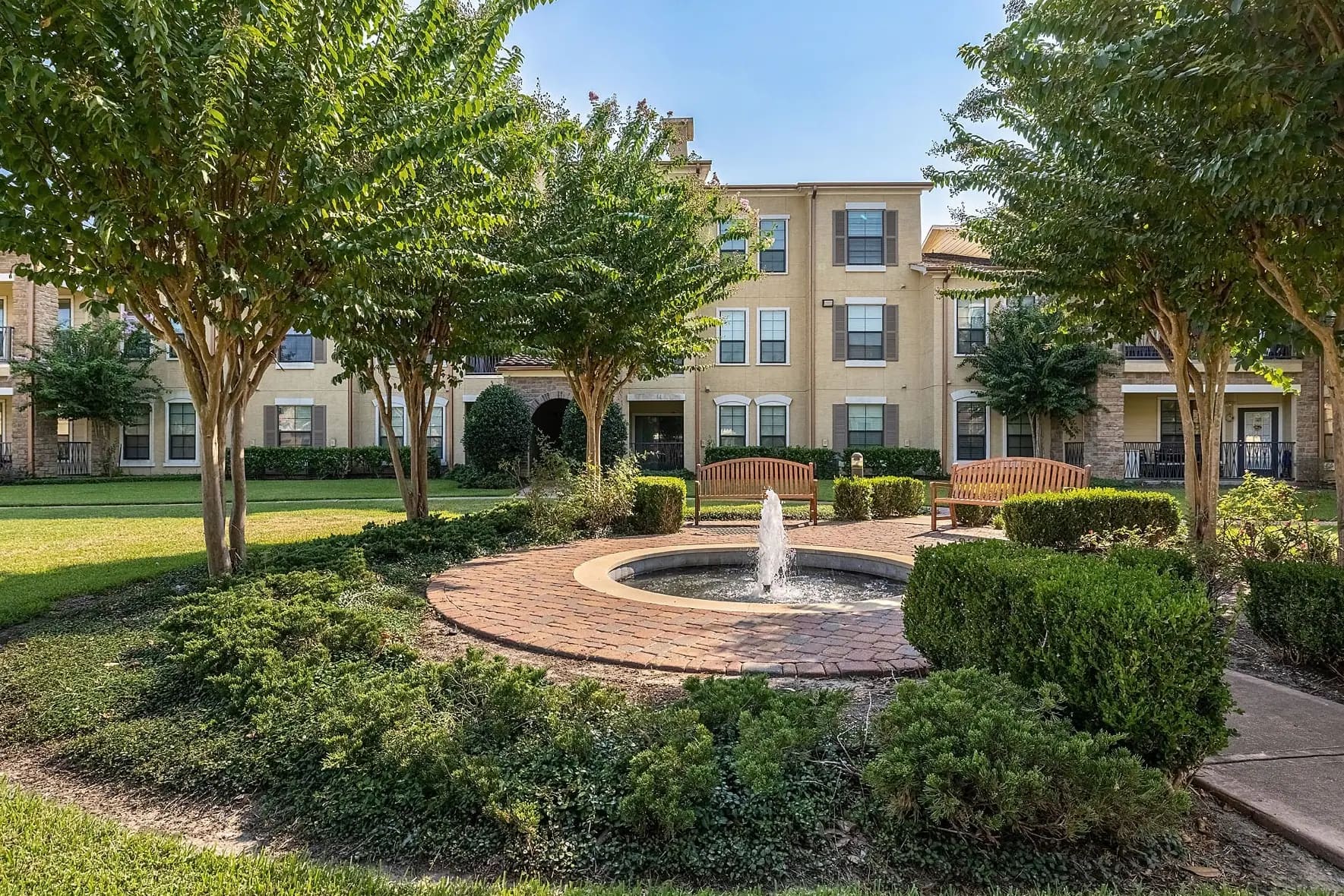 Courtyard with circular brick fountain, mature trees, green hedges, wooden benches, and multi-story residential buildings in background.